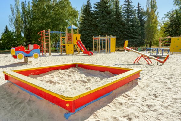 Ein Spielplatz mit einem großen Sandkasten im Vordergrund, dahinter eine Wippe, eine Rutsche und viele Klettermöglichkeiten.