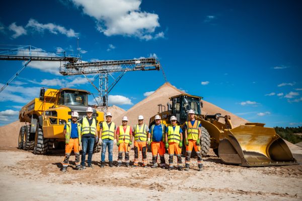 Acht Mitarbeiter des Quarzsandwerkes Wellmersdorf posieren vor einem Muldenkipper und einem Bagger. Im Hintergrund läuft ein Förderband das Quarzsand auf einen großen Sandhügel aufschüttet.