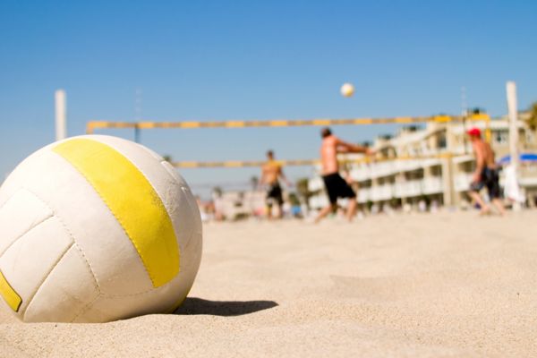 Im Vordergrund liegt ein Volleyball, dahinter ist ein Beachvolleyball auf dem zwei Mannschaften gegeneinander spielen.