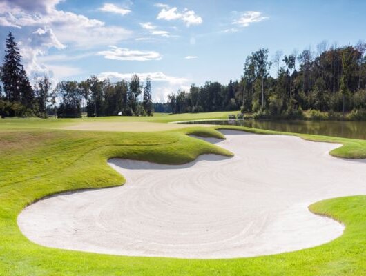 Well-maintained golf course featuring a large white sand bunker beside a green lawn and a pond in the background.