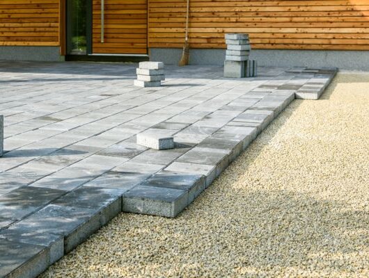 Partially laid gray concrete slabs forming a new terrace beside a wooden house; gravel visible on the side.