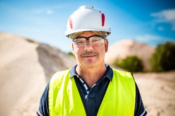 A worker wearing a helmet, goggles, and a yellow vest stands in front of sand mounds, facing the camera.