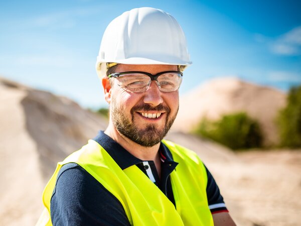 A man wearing a safety helmet, goggles, and a yellow vest smiles in a sunny sand pit.