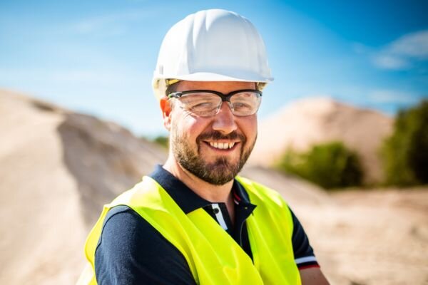 A man wearing a safety helmet, goggles, and a yellow vest smiles in a sunny sand pit.