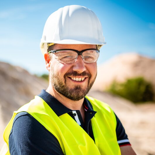 A man wearing a safety helmet, goggles, and a yellow vest smiles in a sunny sand pit.