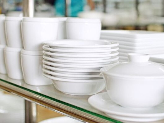Stacked white porcelain dishes with bowls, cups, and plates on a glass shelf in a kitchen or cafeteria.