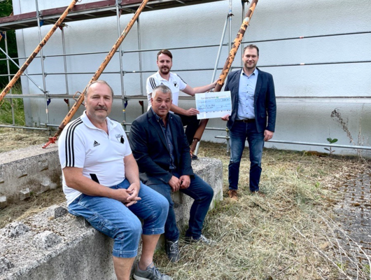 Four men sit and stand in front of a construction site; one symbolically presents a large donation check.