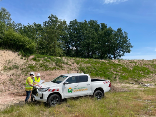 Two people in safety gear stand beside a white company pickup labeled “Wellmersdorfer Quarzsand,” reviewing documents at a sand site.