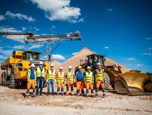 Group of workers in safety gear standing in front of sand piles, construction vehicles, and a conveyor belt.