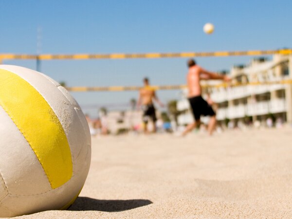 A volleyball lies on the sandy ground while people play beach volleyball in the sunny background.