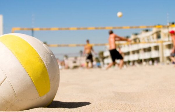 A volleyball lies on the sandy ground while people play beach volleyball in the sunny background.