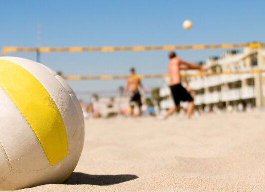 A volleyball lies on the sandy ground while people play beach volleyball in the sunny background.