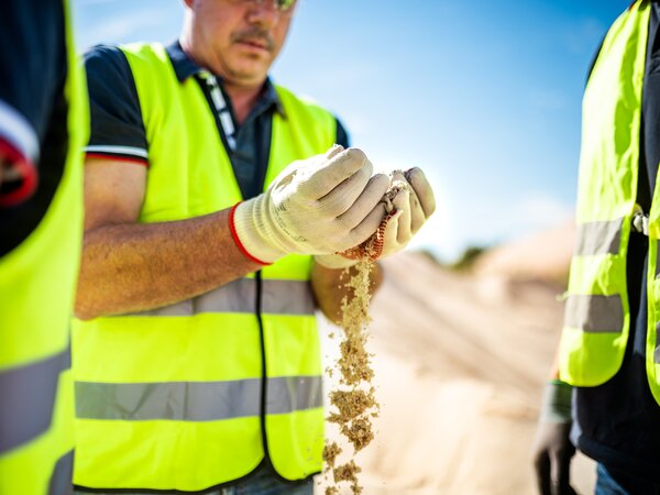 A worker in a safety vest and gloves lets sand run through his hands while inspecting the material.